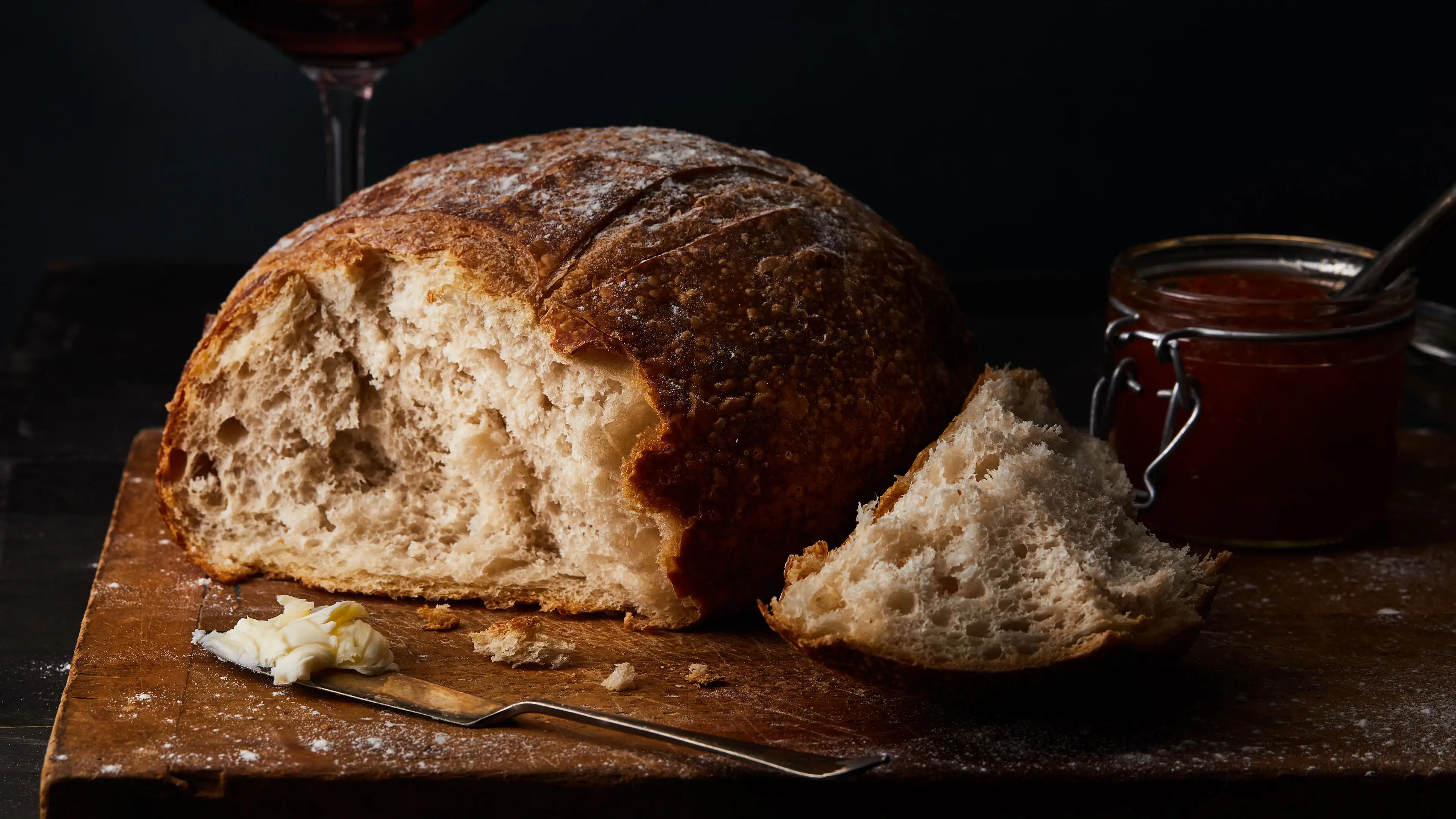 A rustic loaf of bread with a slice cut, rests on a wooden board next to a knife with butter, a jar of jam, and a glass of red wine, all set against a dark background.