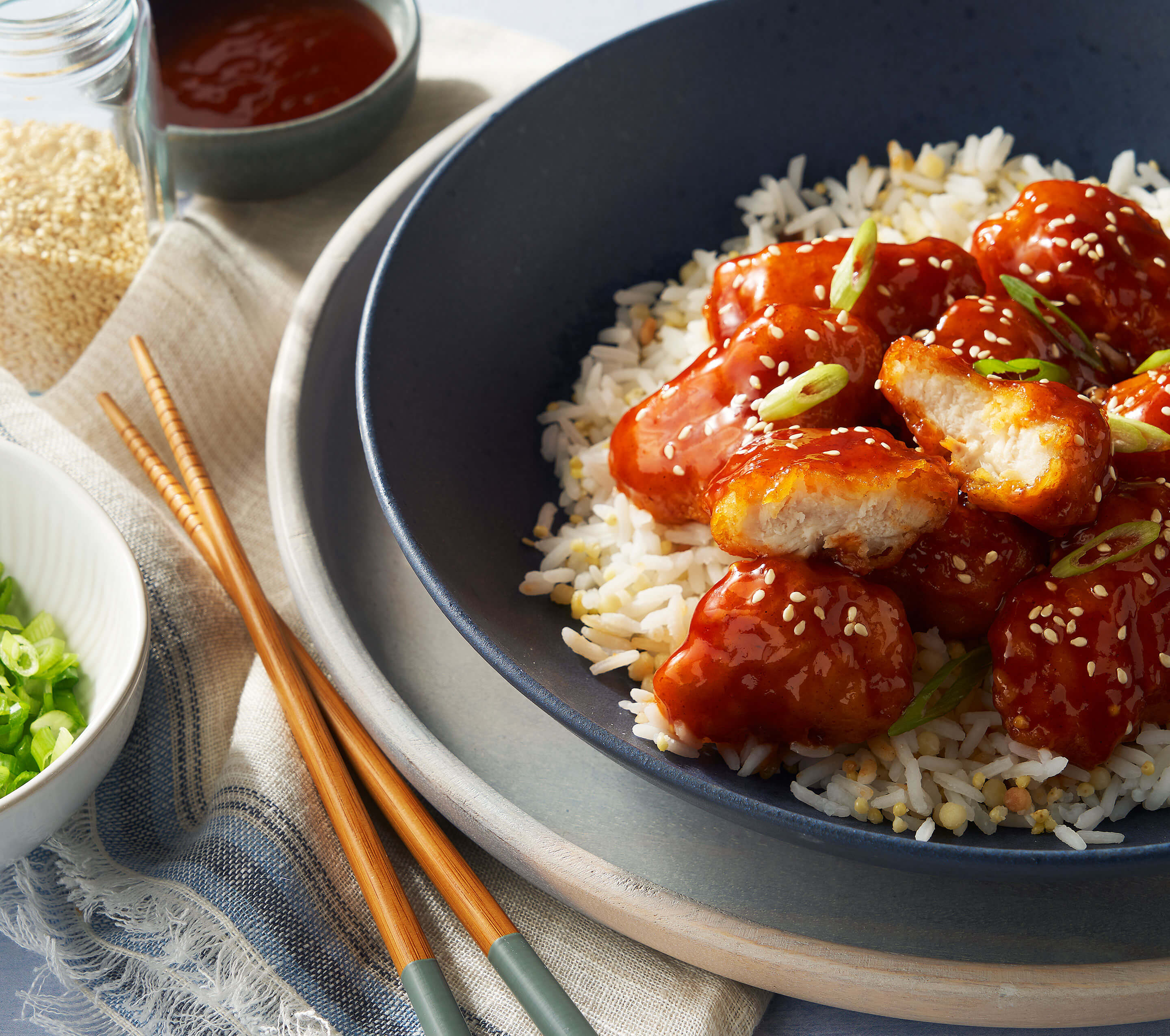 A bowl of white rice topped with saucy sesame chicken pieces, garnished with sliced green onions and sesame seeds. Chopsticks rest nearby, with a small bowl of green onions and a jar of sauce in the background.