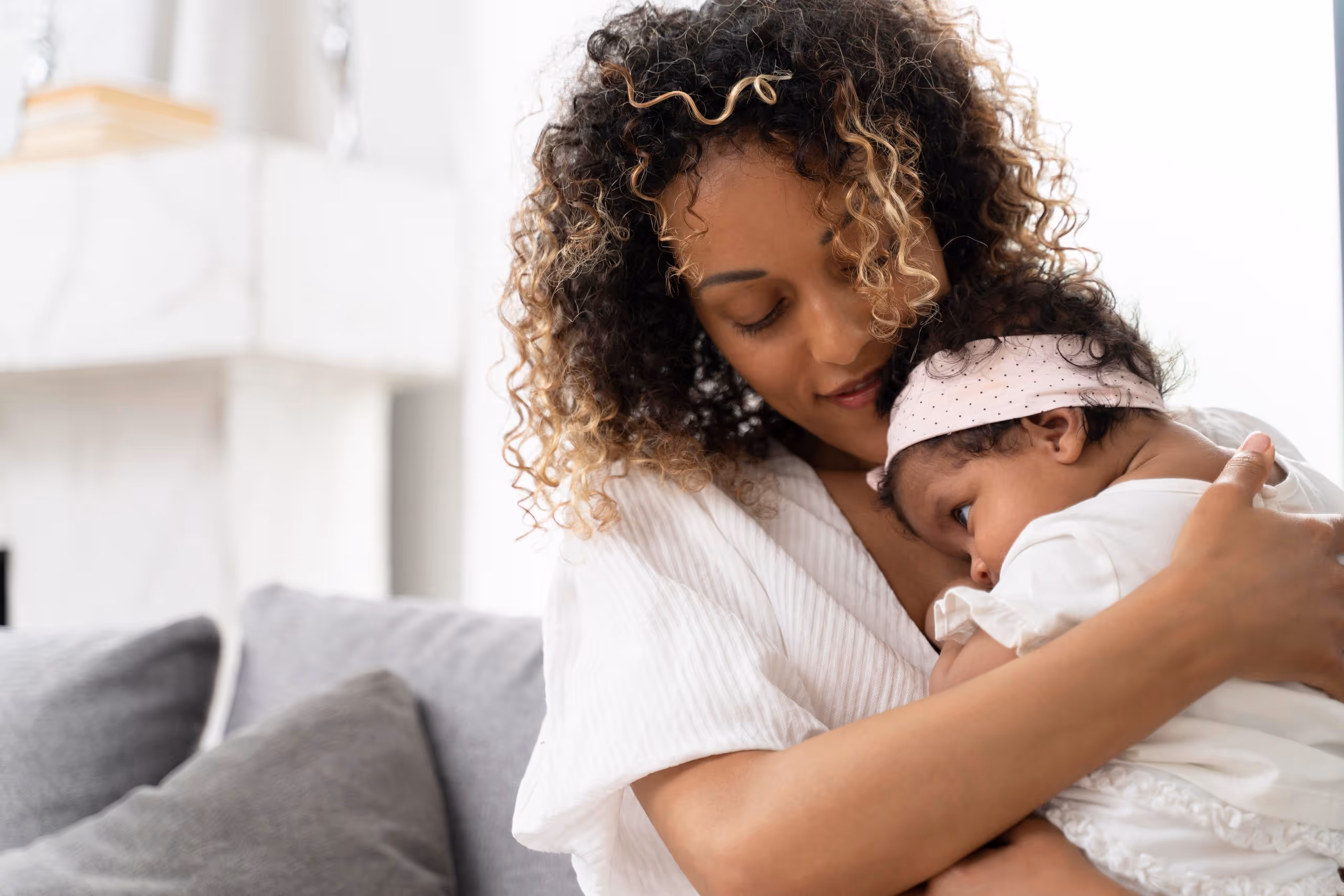 A woman gently embraces a baby, creating a tender and loving atmosphere. The cozy setting features a couch and soft lighting, enhancing the warmth.