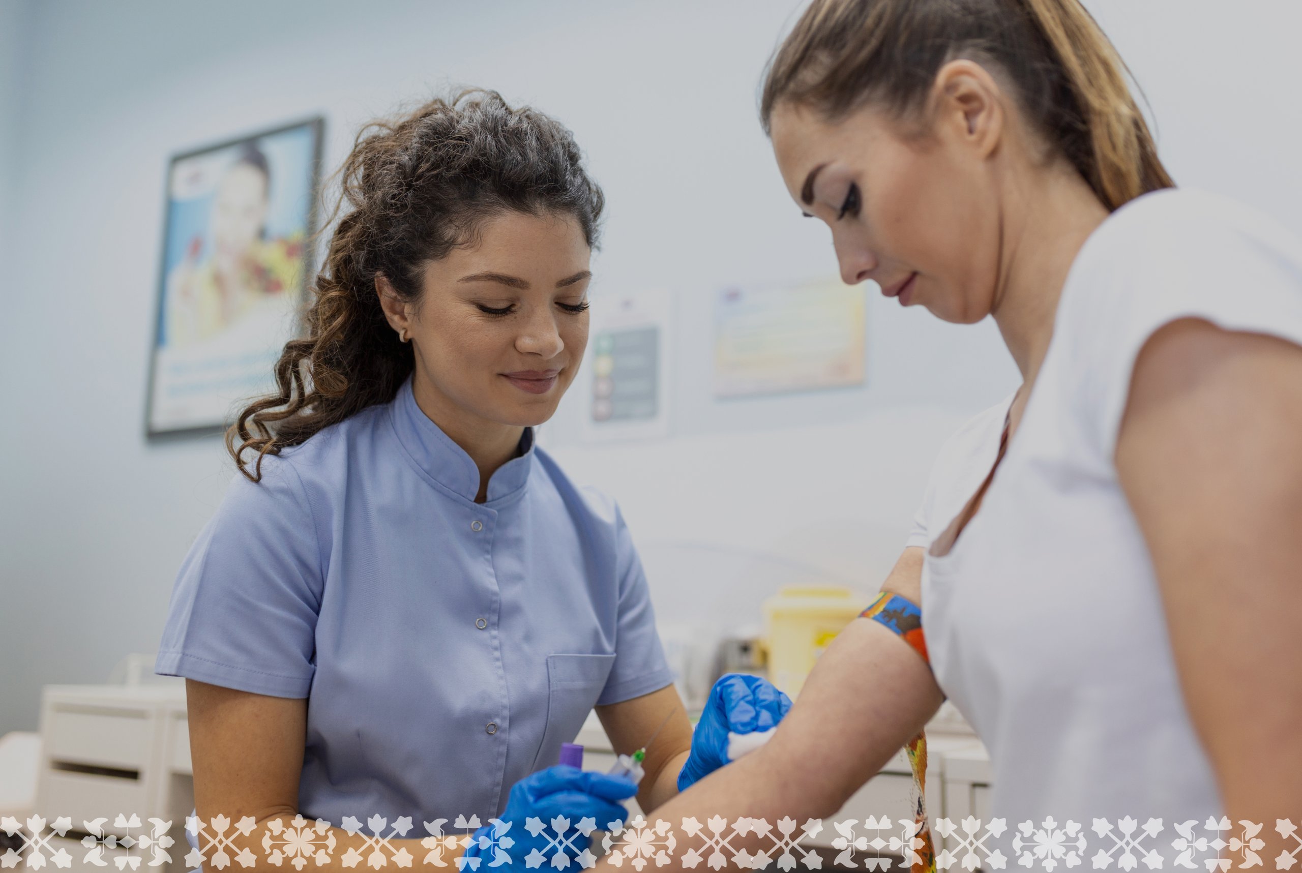 A nurse in a light blue uniform attentively draws blood from a patient wearing a white top. The setting is a clean, clinical room with posters on the wall.