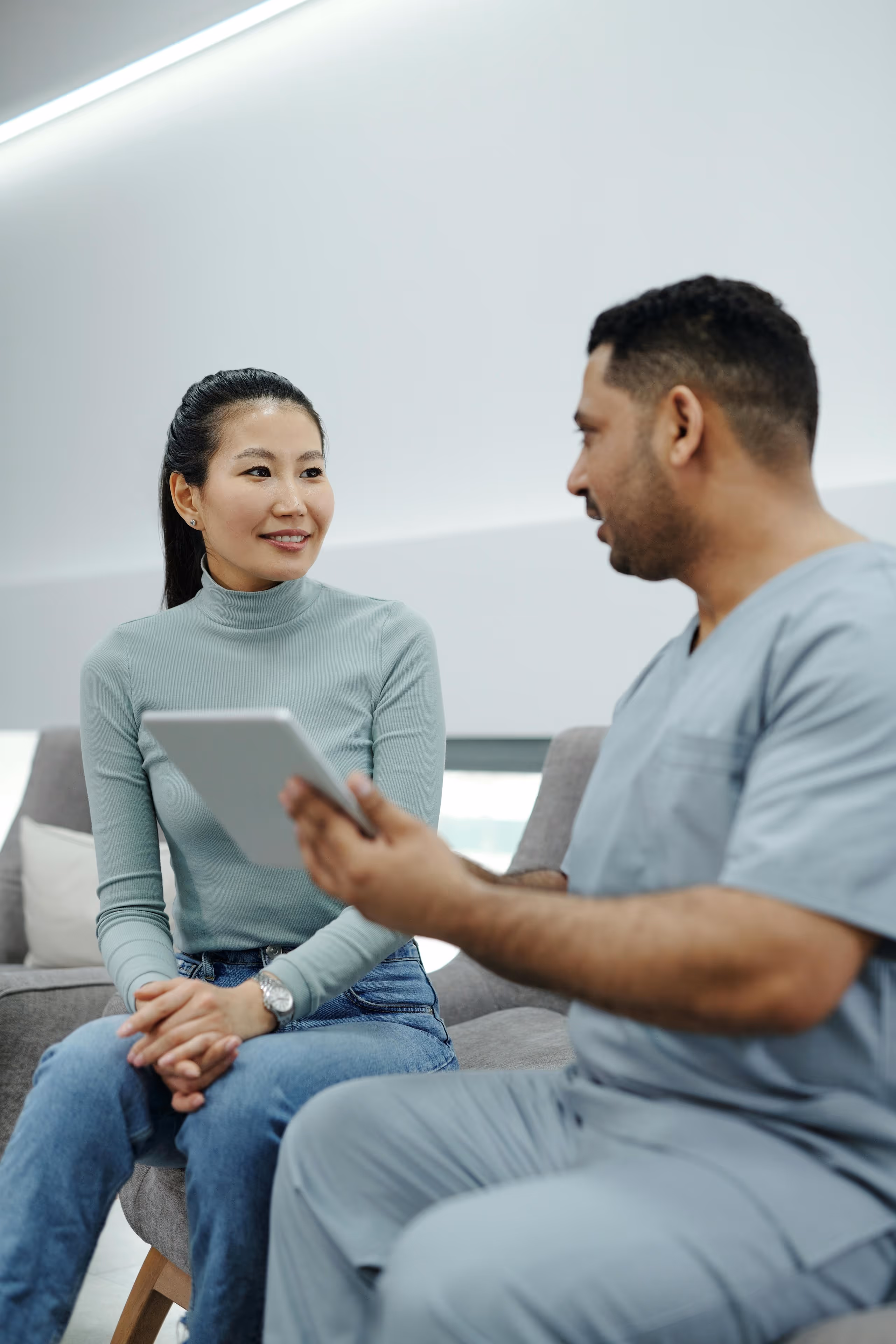 A healthcare professional in scrubs discusses with a seated woman holding a tablet. They sit in a modern, bright room, conveying a calm, professional tone.