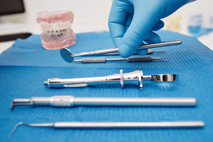 Gloved hand selects dental tool from a set on a blue tray. A model of teeth is blurred in the background, suggesting a clinical setting.