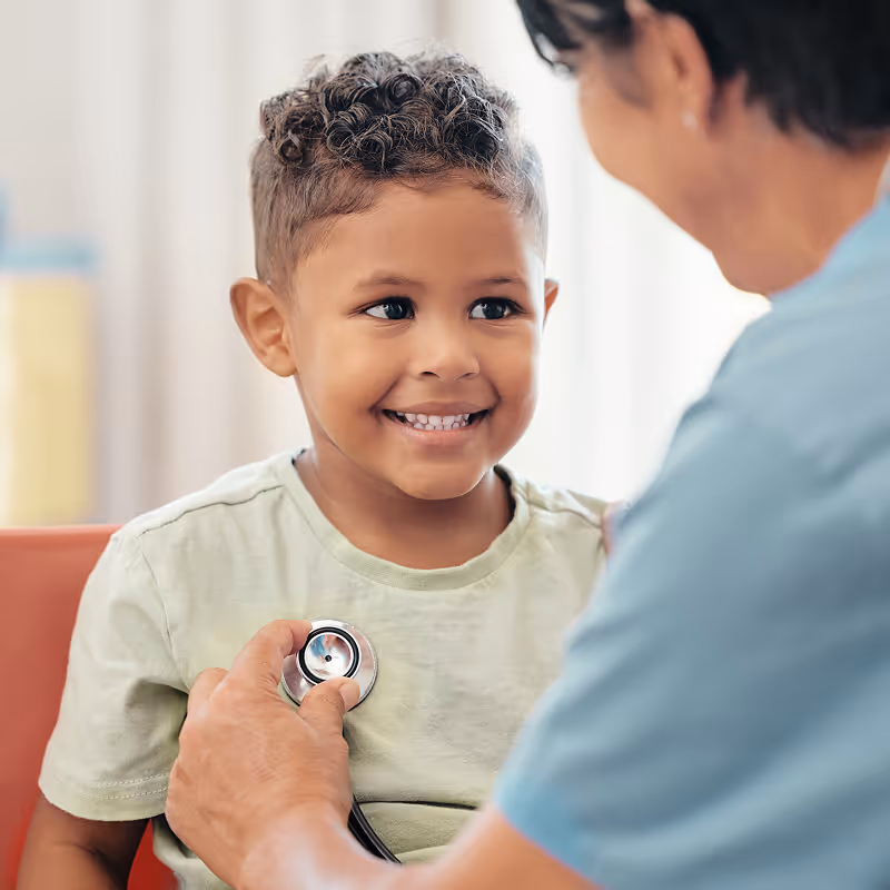 Young boy smiling as a healthcare professional in blue scrubs holds a stethoscope to his chest. The setting is a bright, comforting medical office.