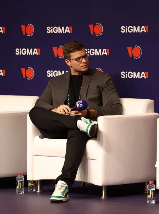 Benedikt Becker sitting on a panel stage at SiGMA, holding a microphone and listening during a discussion.