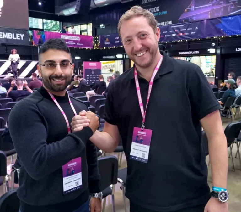 Two men smiling and shaking hands at a conference, wearing event badges with a seated audience in the background.