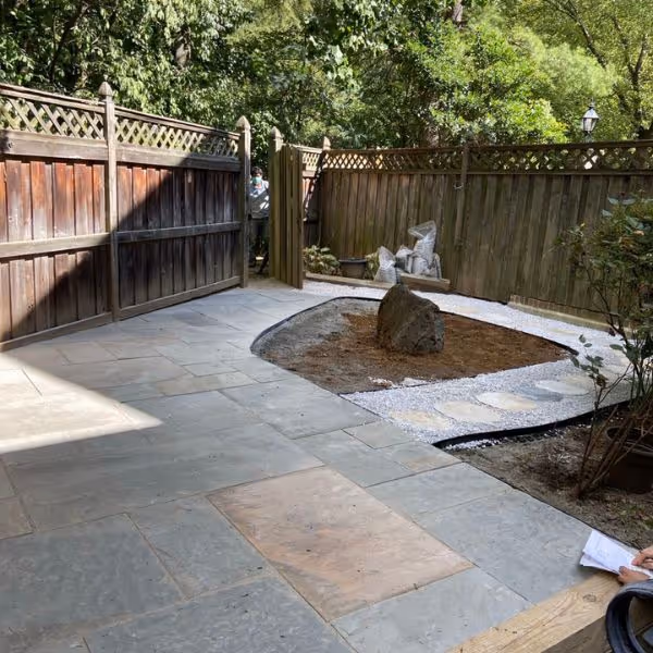 Backyard patio with stone paving, a fenced wooden gate open to a garden area with a central large rock, surrounded by trees.