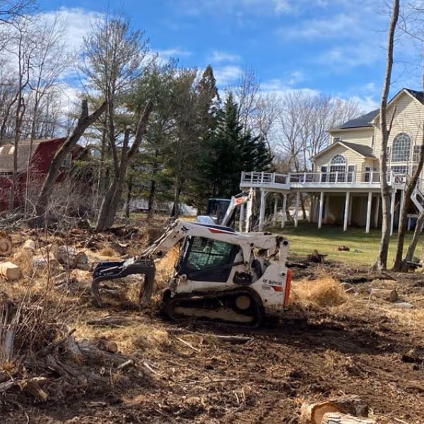 Compact excavator clearing tree stumps and debris in a yard near a large house with a deck.