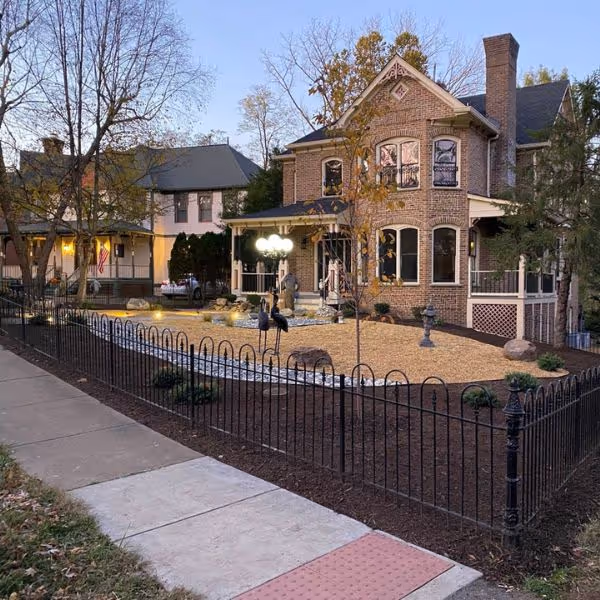 Two historic houses with front yards featuring manicured landscaping including a lawn with boulders, lamp posts, and small trees, bordered by a black wrought iron fence.