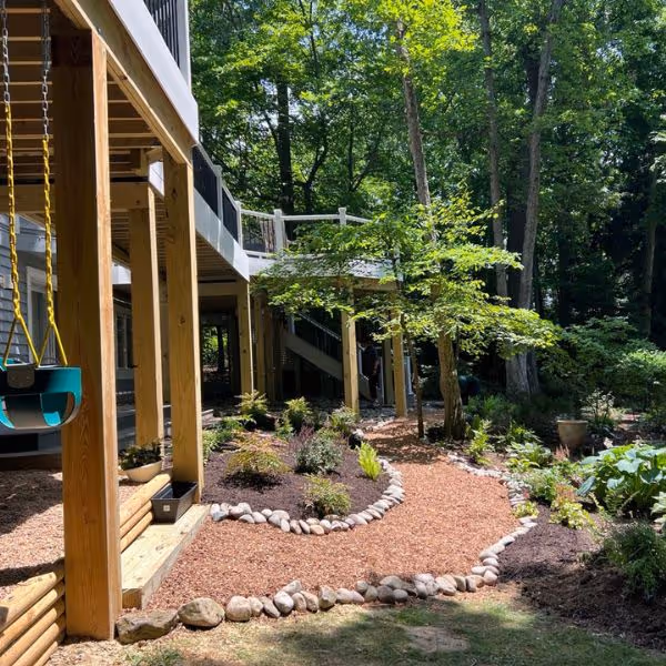 Garden path with reddish mulch bordered by stones, surrounded by plants and trees next to a wooden deck with children's swings.