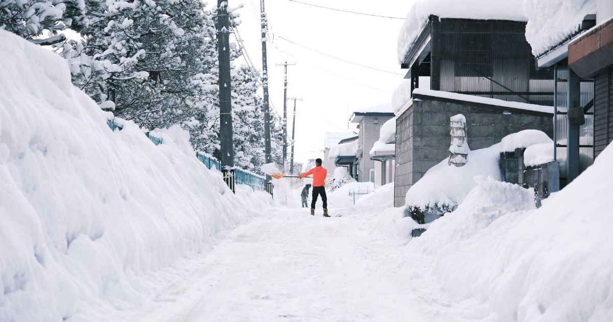 Person in red jacket shoveling snow on a narrow street between houses