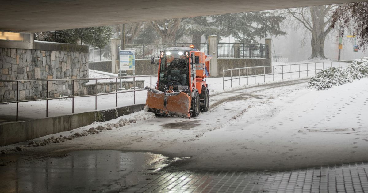 Orange snow plow clearing snowy walkway near stone walls and trees