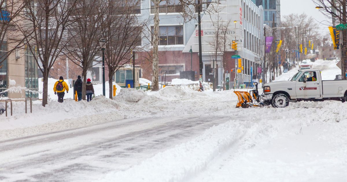 Snow-covered urban street with snowplow and pedestrians walking on cleared sidewalk
