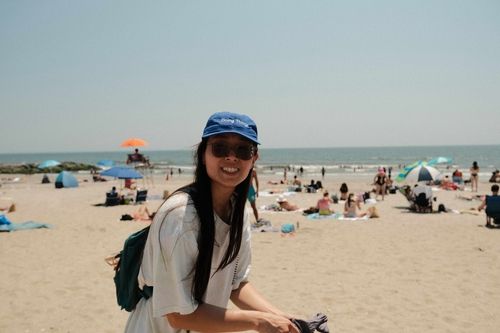 Photo of Giovanna at the beach. She's wearing a blue cap that says "Doing Things" on the front. She's wearings sunglasses and smiling directly at the camera. She's wearing an oversized light colored t-shirt and a backpack.
