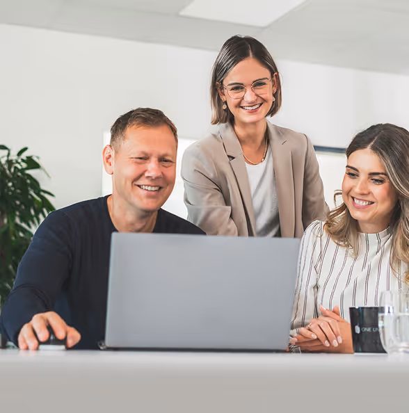 Drei lächelnde Personen, zwei Frauen und ein Mann, schauen gemeinsam auf einen Laptop in einem modernen Büro.