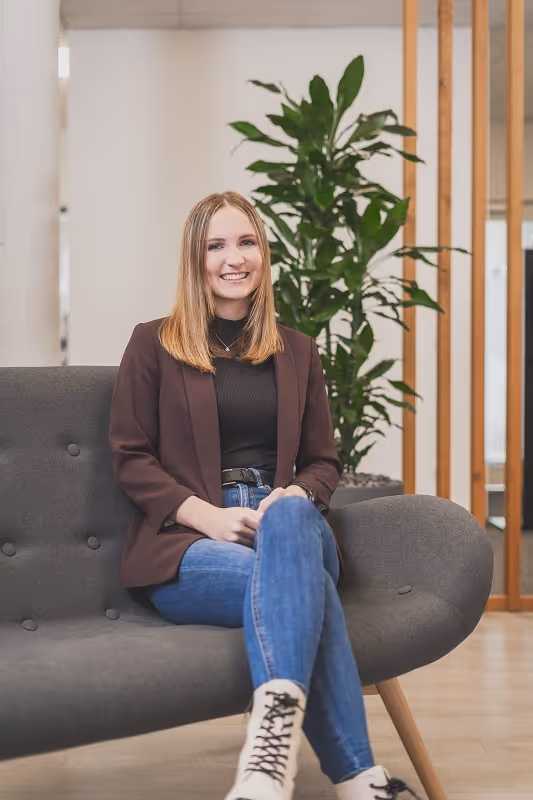 Lächelnde Frau mit braunem Blazer, schwarzem Oberteil und blauen Jeans sitzt auf grauem Sofa vor grüner Pflanze in modernem Raum.