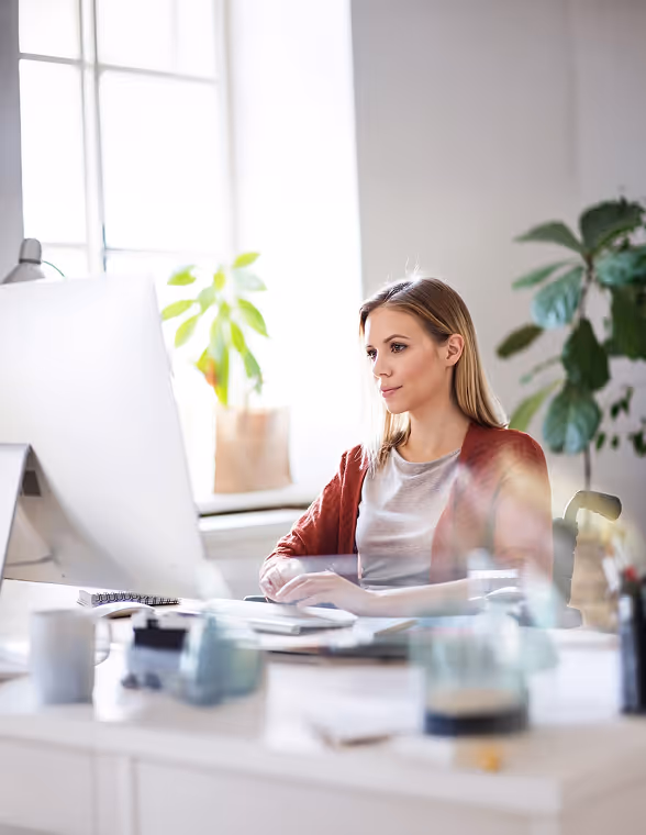 Frau arbeitet konzentriert an einem Computer in einem hellen Büro mit Pflanzen im Hintergrund.