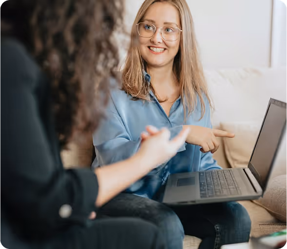 Junge Frau mit Brille zeigt lächelnd auf Laptop-Bildschirm, während sie mit einer anderen Person spricht.