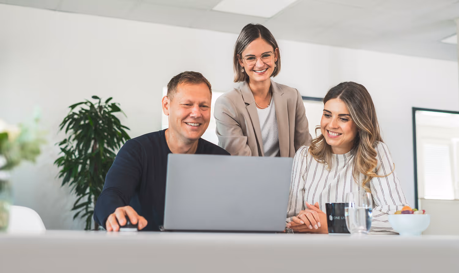 Drei lächelnde Mitarbeiter arbeiten gemeinsam an einem Laptop in einem modernen Büro.
