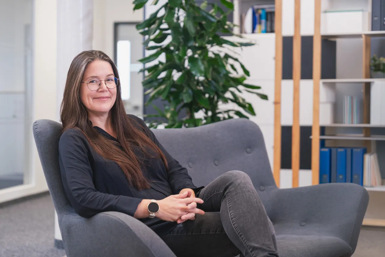 Frau mit langen braunen Haaren und Brille sitzt lächelnd auf einem grauen Sofa in einem modernen Büro.