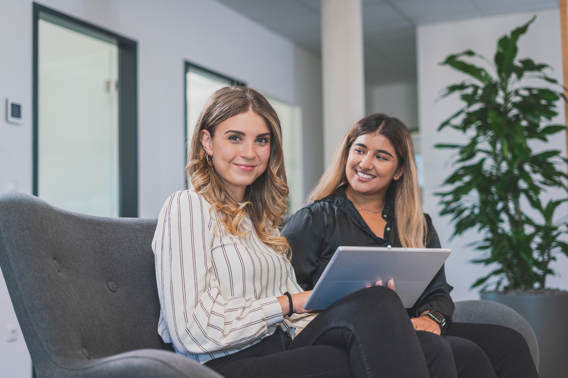 Zwei lächelnde Frauen sitzen auf einer grauen Couch, eine hält ein Tablet, im modernen Büro mit großen Pflanzen im Hintergrund.