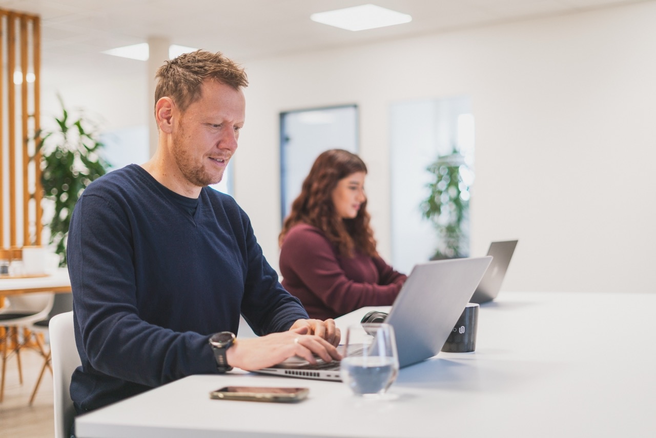 Mann und Frau arbeiten an Laptops an einem weißen Tisch in einem modernen Büro mit Pflanzen im Hintergrund.