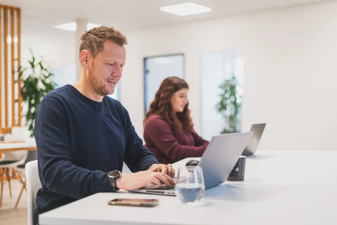 Mann und Frau arbeiten an Laptops an einem weißen Tisch in einem modernen Büro mit Pflanzen im Hintergrund.