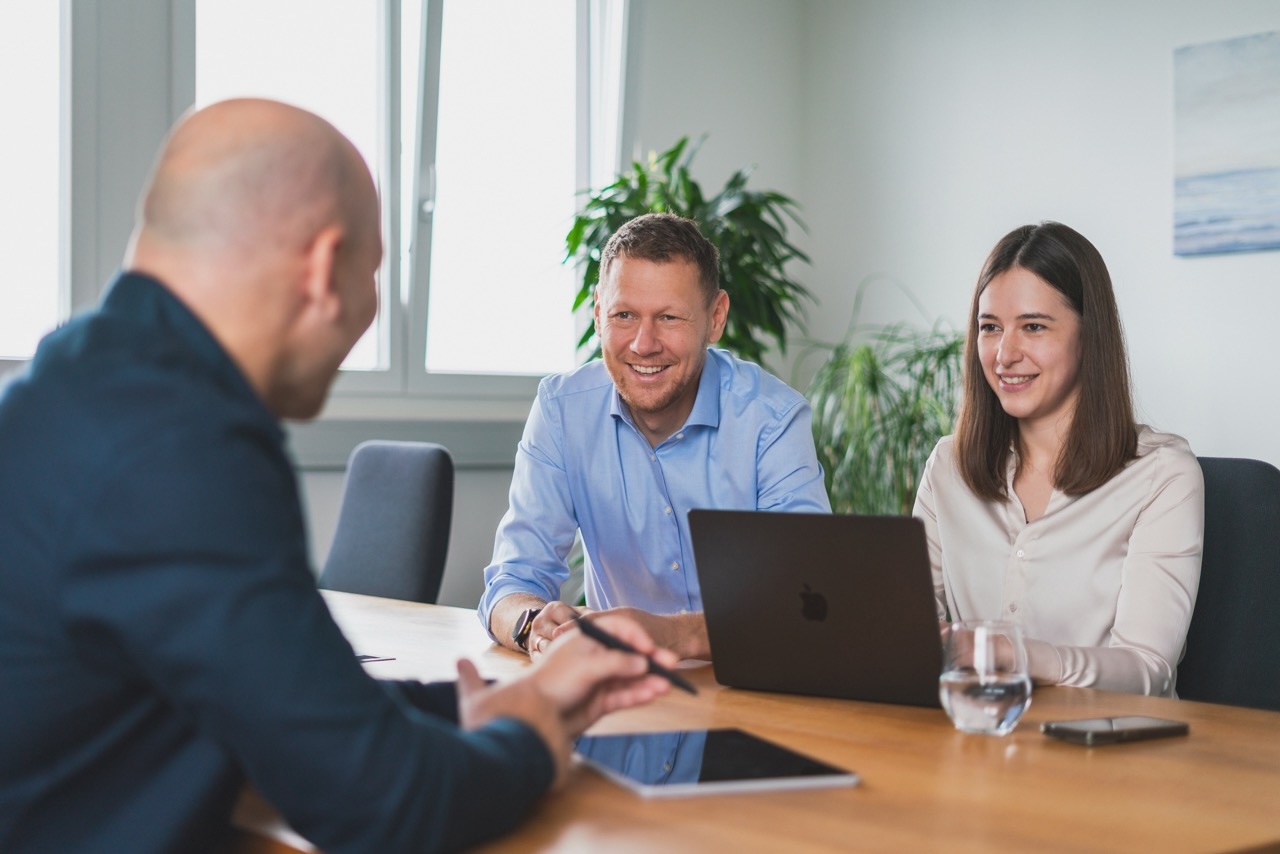 Drei Personen sitzen an einem Konferenztisch und führen ein freundliches Gespräch in einem Büro.