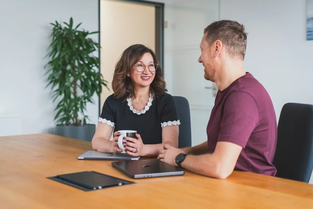 Zwei lächelnde Personen, eine Frau mit Tasse und ein Mann, sitzen an einem Holztisch mit Laptops in einem modernen Büro.