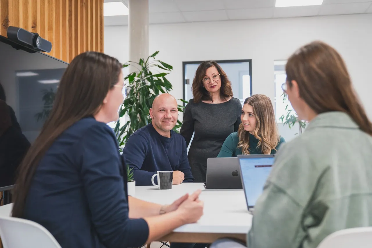 Fünf Personen in einem modernen Büro sitzen um einen Tisch und diskutieren während eines Meetings mit Laptops und Tassen.