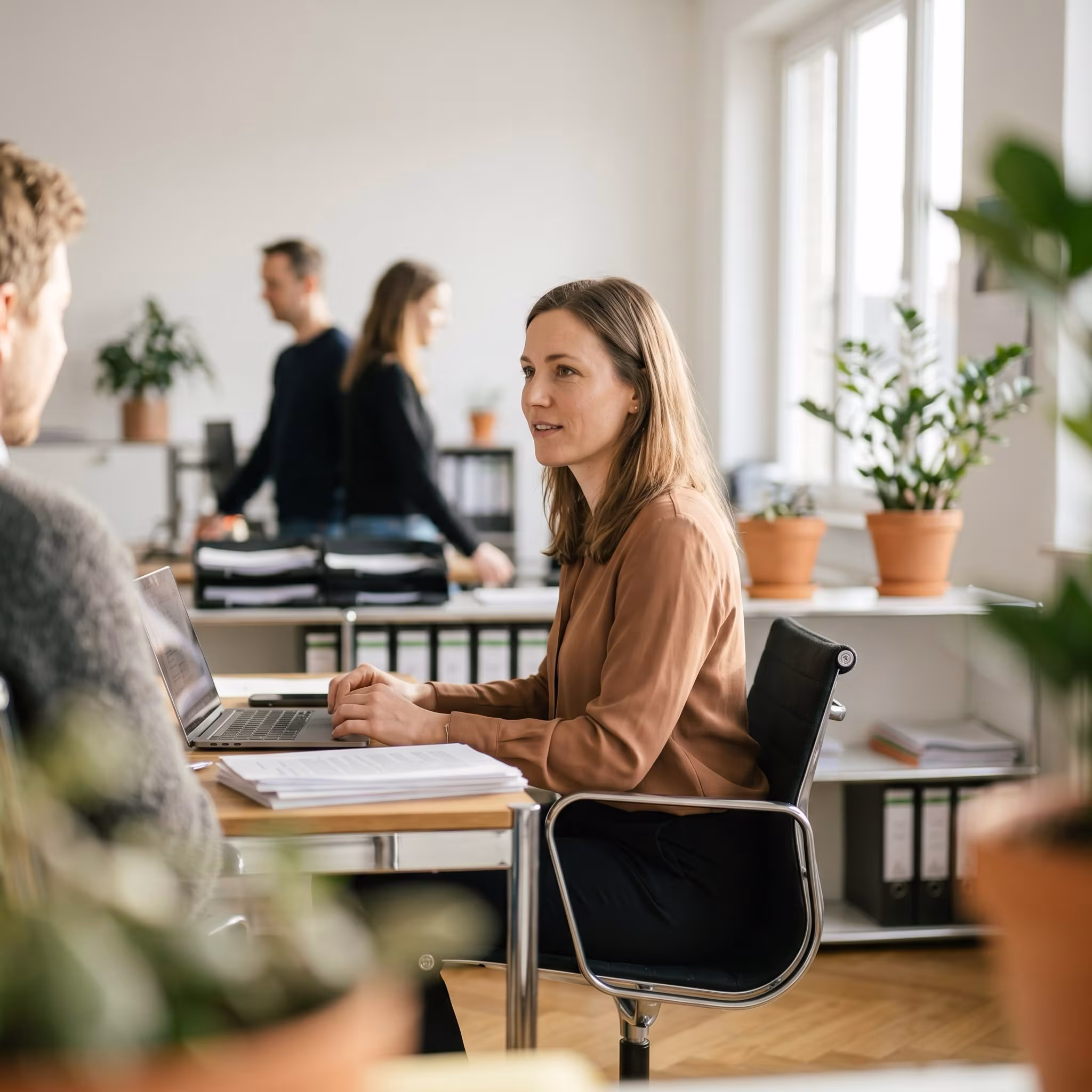 Frau sitzt an einem Schreibtisch und unterhält sich mit einem Mann in einem Büro mit Pflanzen und aktenschränken.