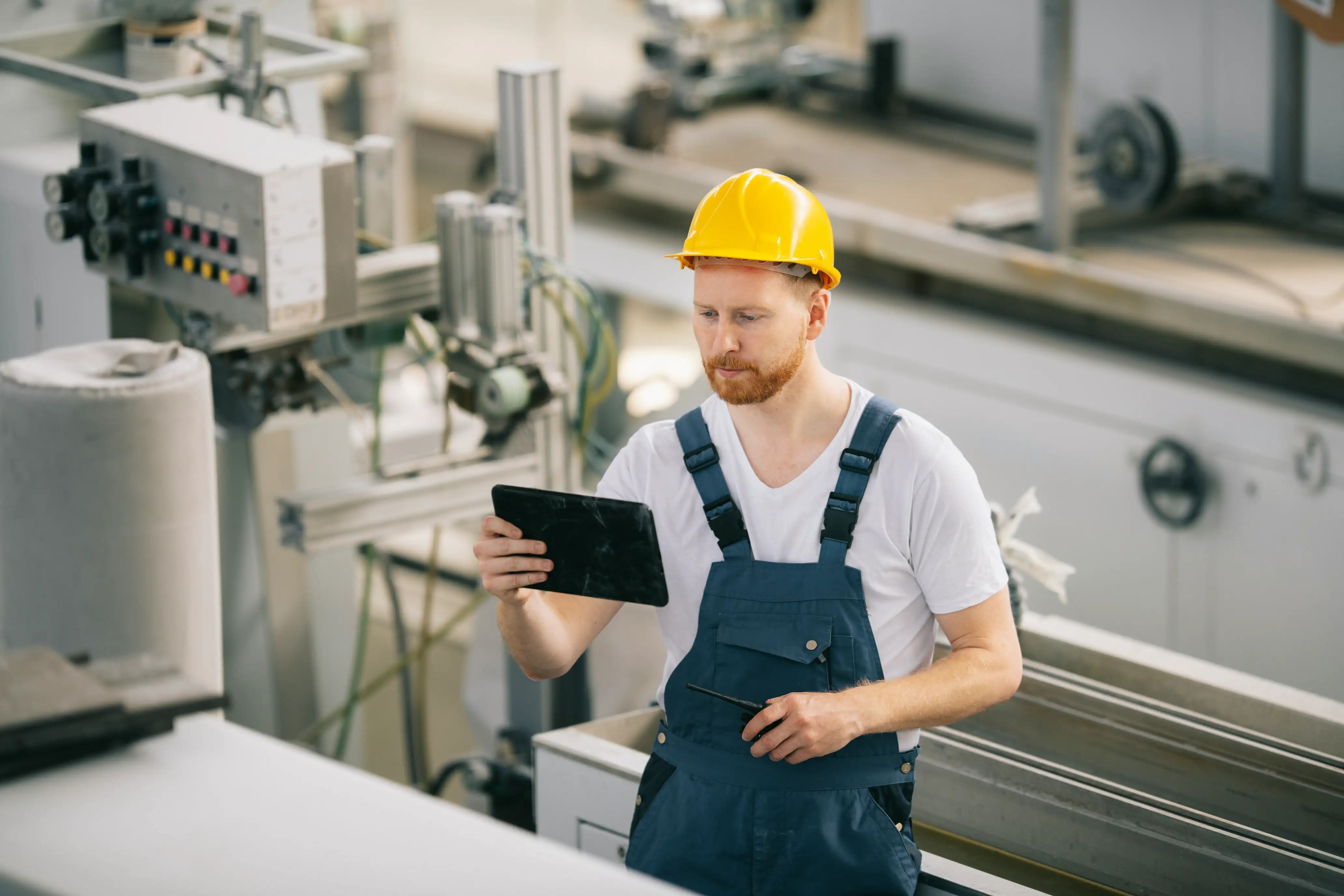 Industriearbeiter mit gelbem Helm und blauer Arbeitskleidung, der in einer Fabrikhalle ein Tablet betrachtet.