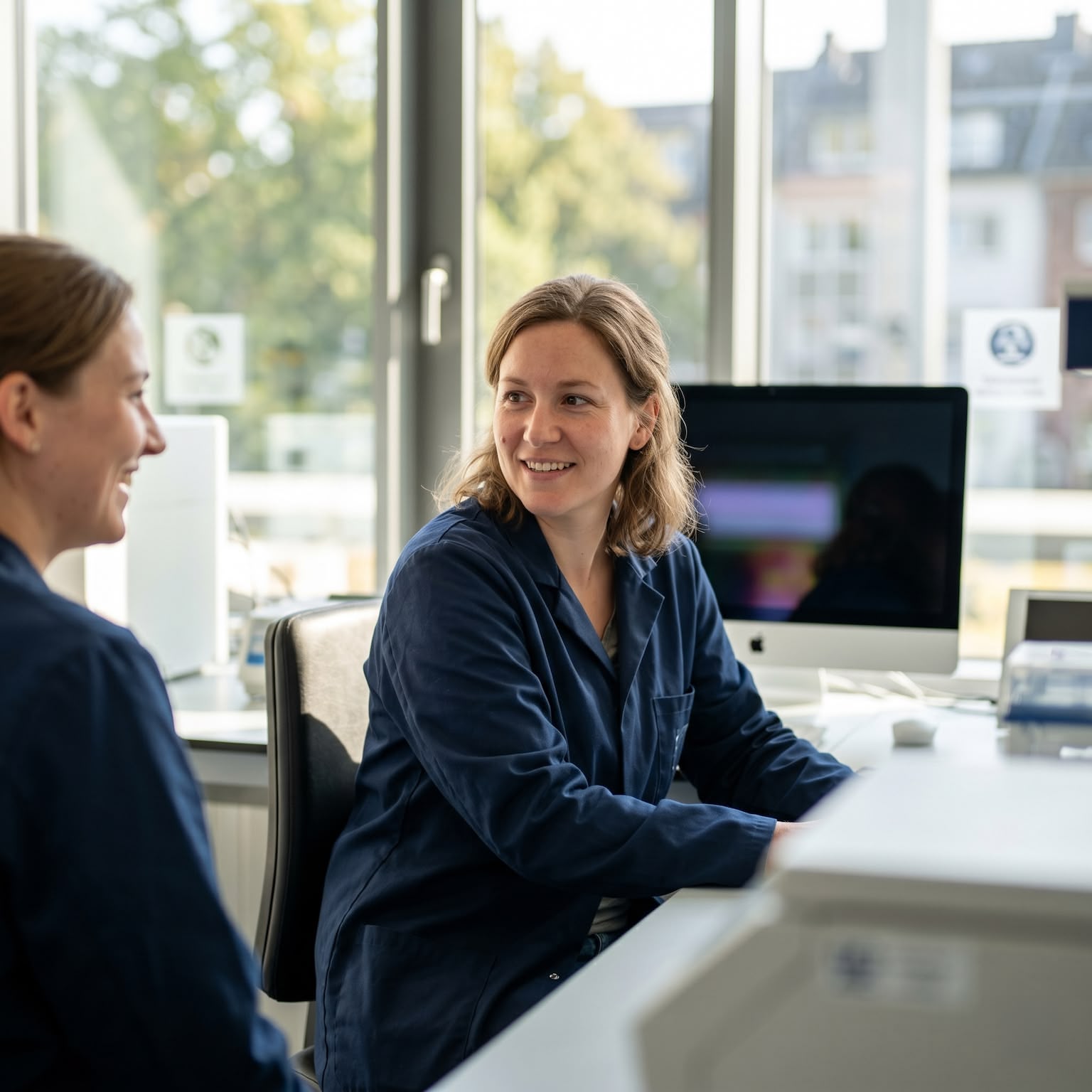 Zwei Frauen in blauen Laborkitteln unterhalten sich freundlich in einem hellen Büro mit Computer.