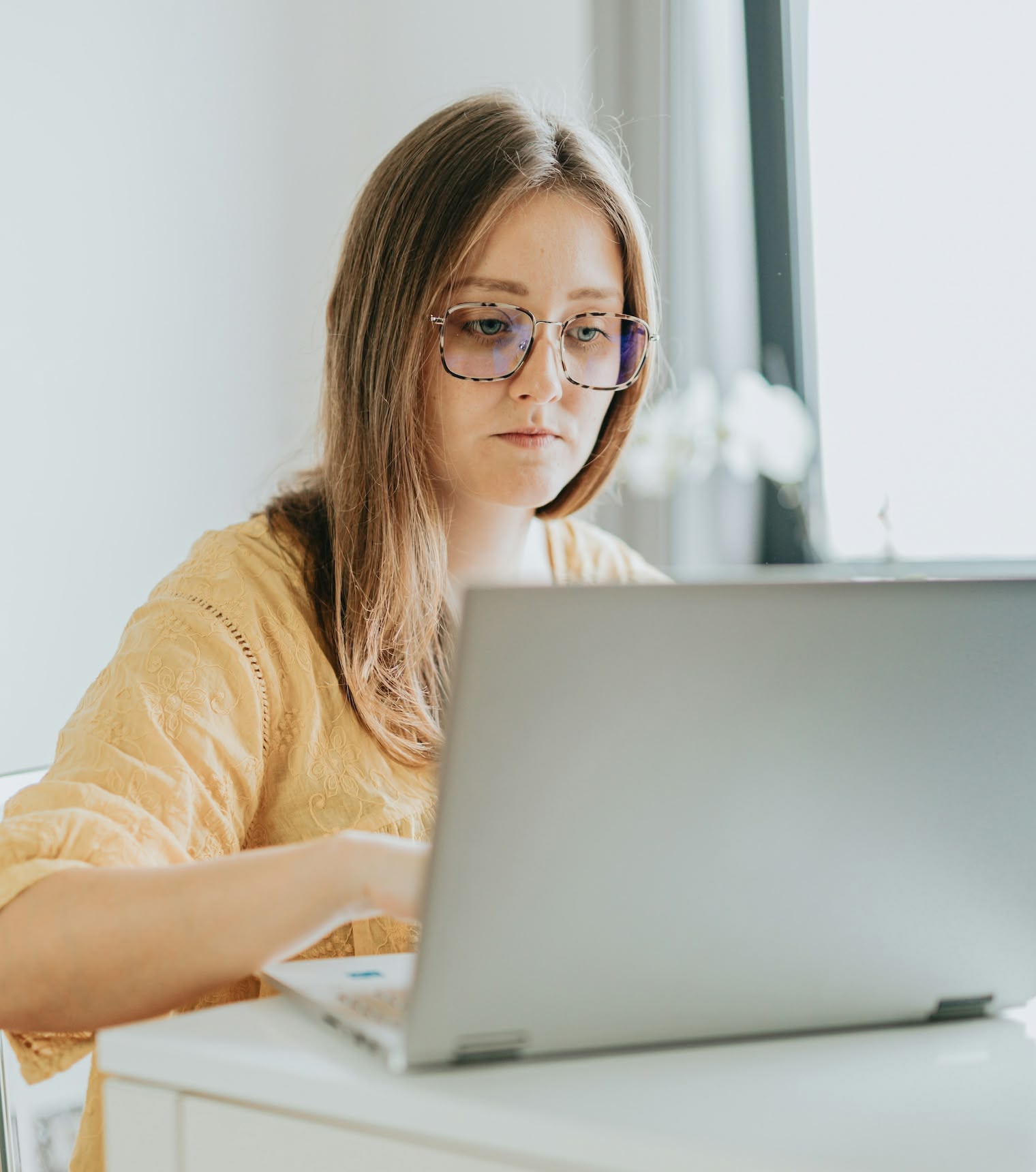 Junge Frau mit Brille arbeitet konzentriert an einem Laptop in einem hellen Raum.