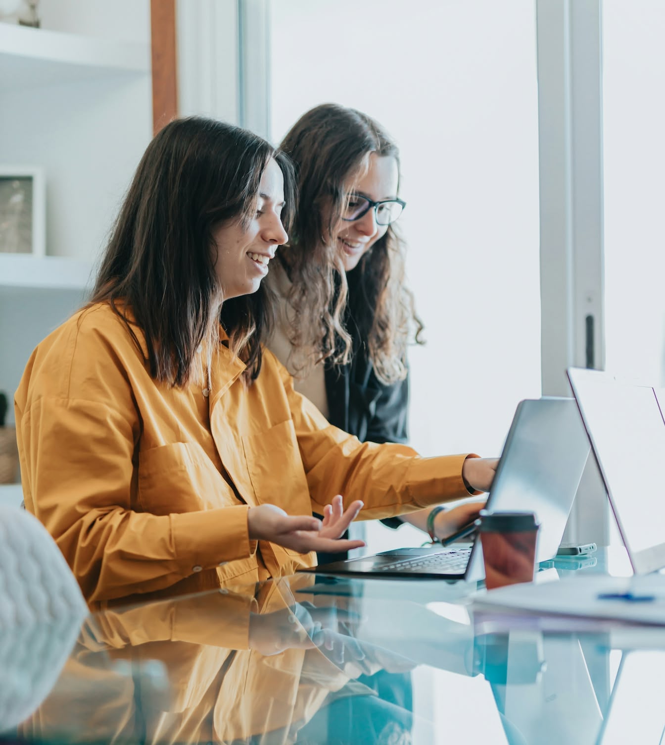 Zwei Frauen im Büro arbeiten gemeinsam an einem Laptop, lächeln und diskutieren.