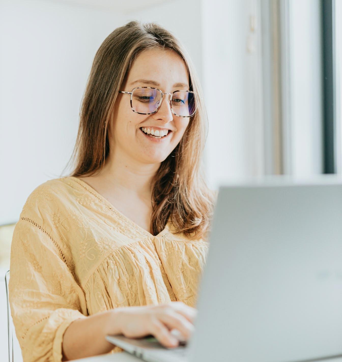 Junge Frau mit Brille und hellgelbem Oberteil, die lächelnd an einem Laptop arbeitet.