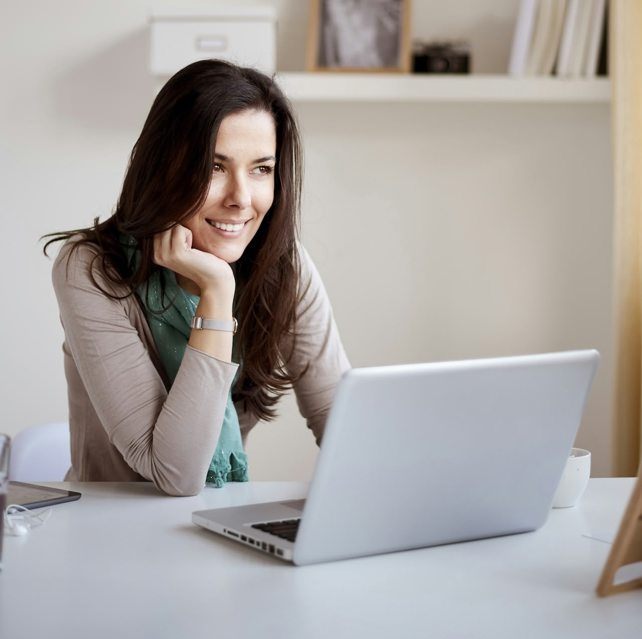 Lächelnde Frau sitzt zu Hause am Tisch und schaut auf einen offenen Laptop.