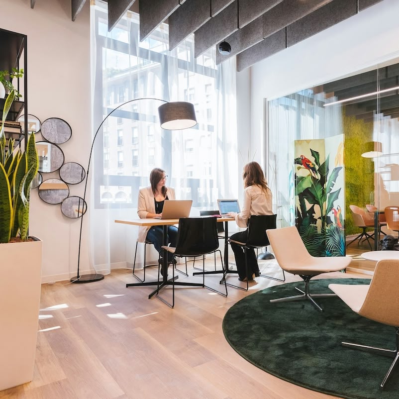 Zwei Frauen arbeiten an Laptops an einem Tisch in einem modernen, lichtdurchfluteten Büro mit Pflanzen und Glaswänden.