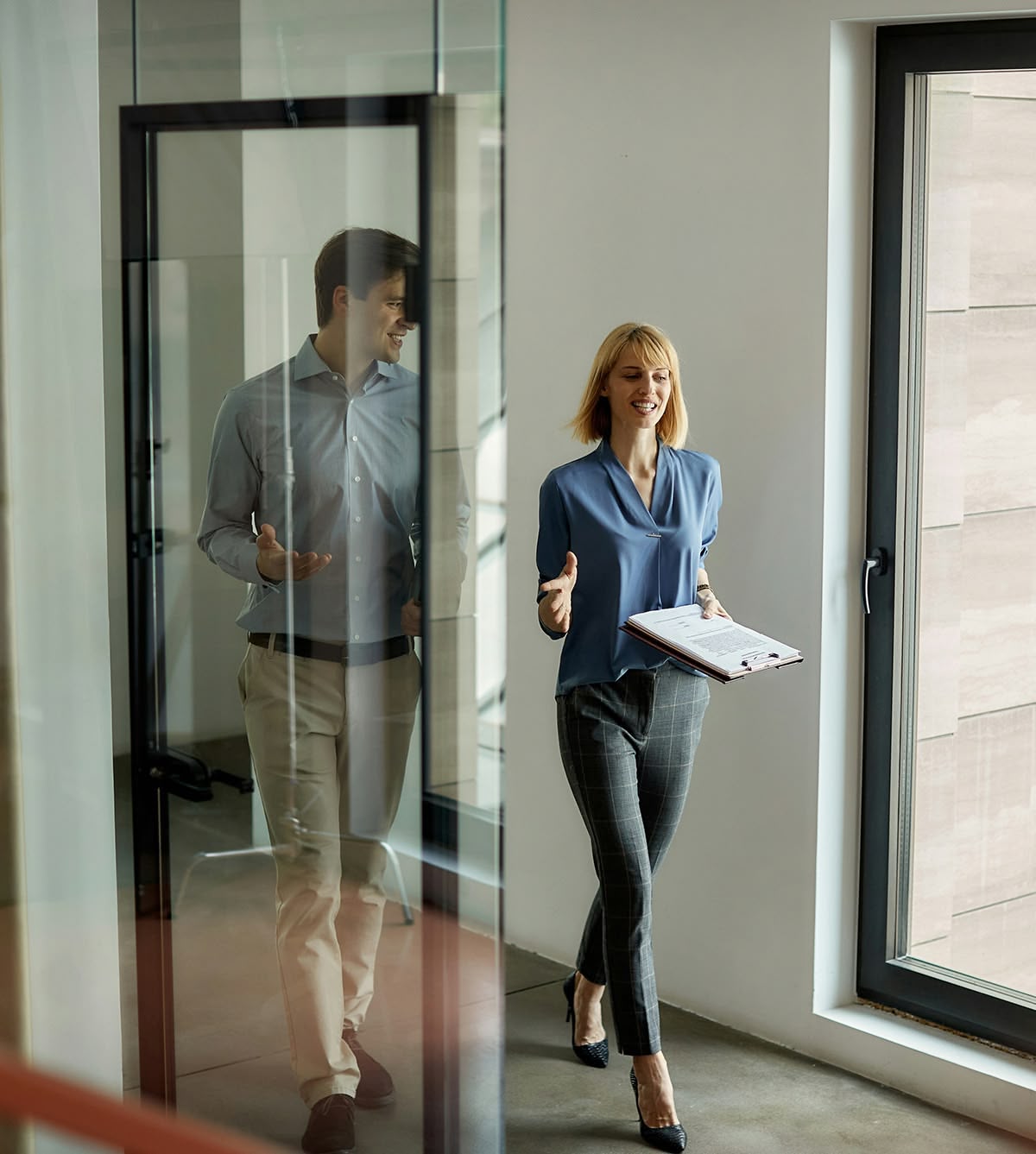Zwei professionelle Büroangestellte gehen und unterhalten sich in einem modernen Büroflur mit Fenster.