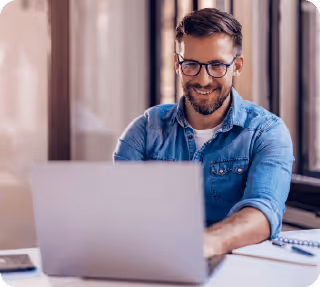 Lächelnder Mann mit Brille und Jeanshemd arbeitet an einem Laptop in einem hellen Raum.