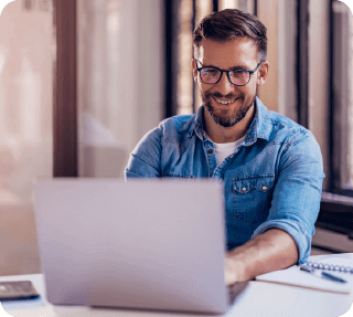 Lächelnder Mann mit Brille und Jeanshemd arbeitet an einem Laptop in einem hellen Raum.