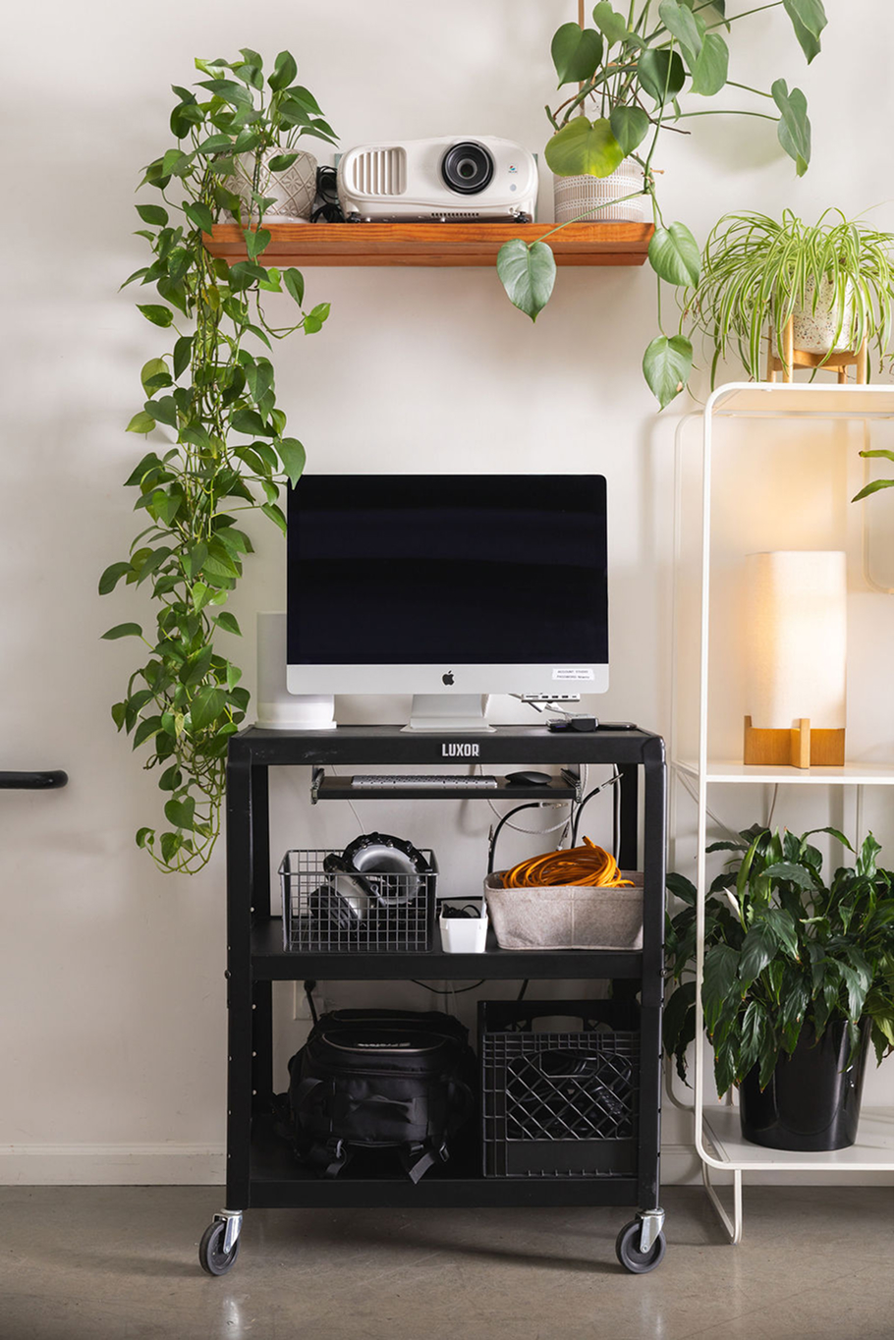 A black cart with wheels displaying a desktop monitor, headphones, and storage bins. Above, a wooden shelf holds a projector, surrounded by green plants. A lighted plant stand is on the right side.