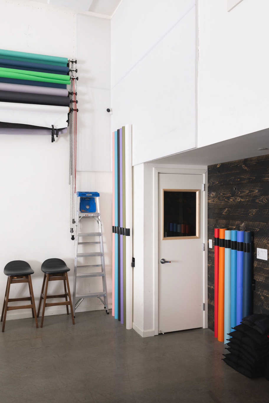 An interior studio space with a ladder, two stools, and colorful background paper rolls in vertical holders. A closed door features a window, and the wall has a dark wood paneling section.