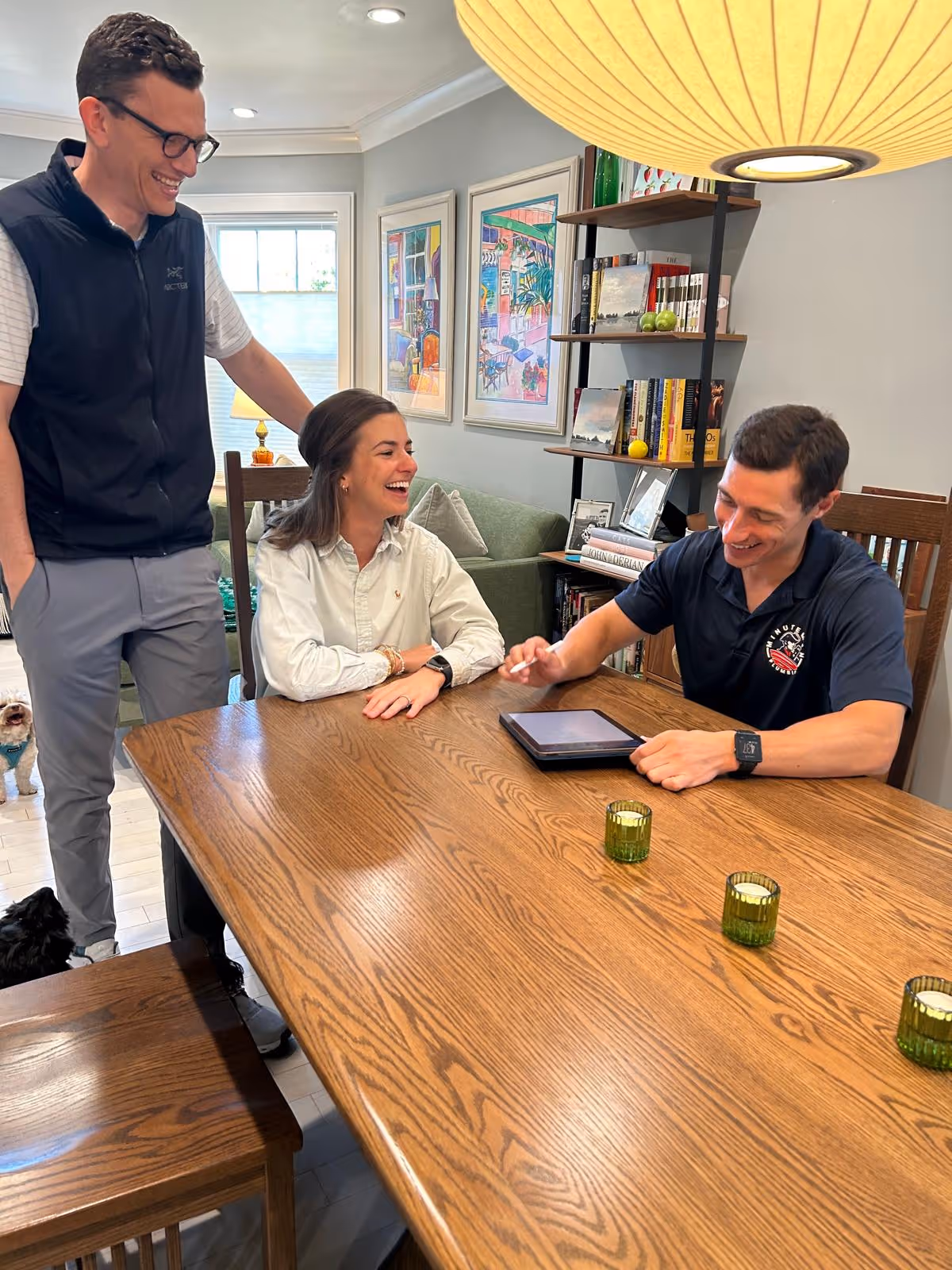 Minuteman Plumbing, Heating & Cooling technician meeting with smiling homeowners at their dining table to review a project estimate on a tablet.