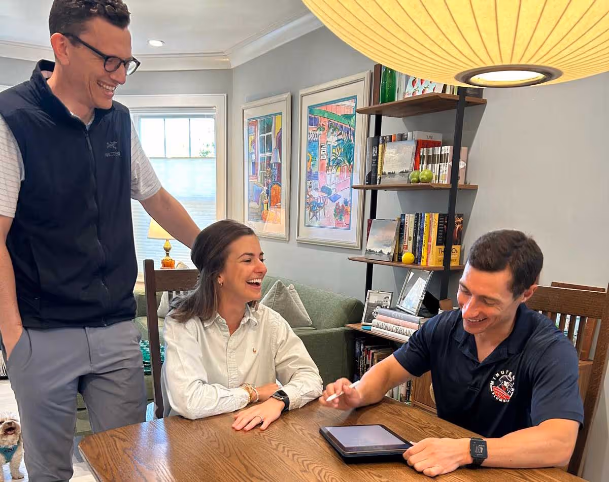 Minuteman Plumbing, Heating & Cooling technician meeting with smiling homeowners at their dining table to review a project estimate on a tablet.