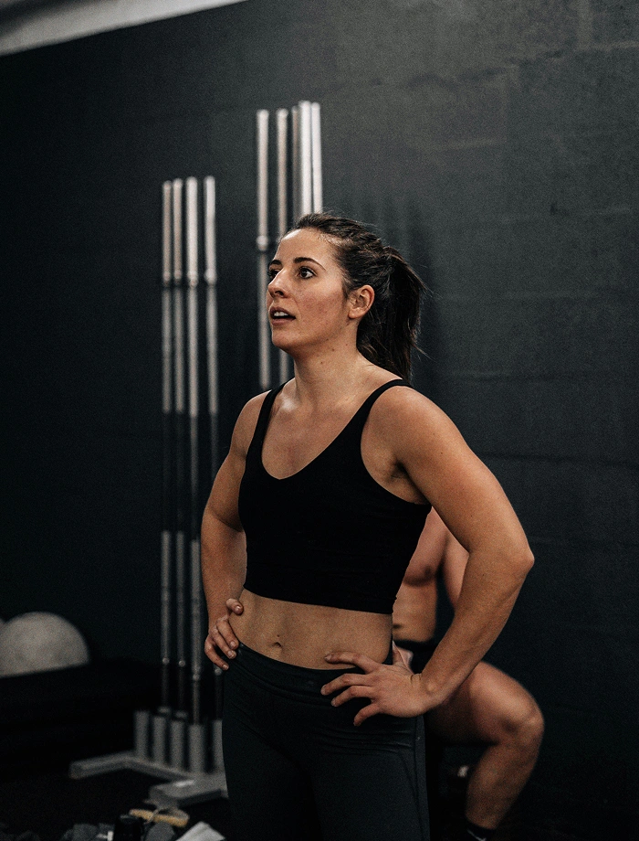 Fit woman in black workout clothes standing with hands on hips in a gym looking focused.
