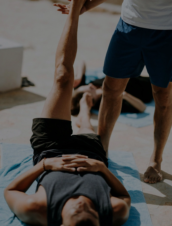 Person lying on a blue mat with one leg raised, assisted by another person standing nearby in blue shorts.