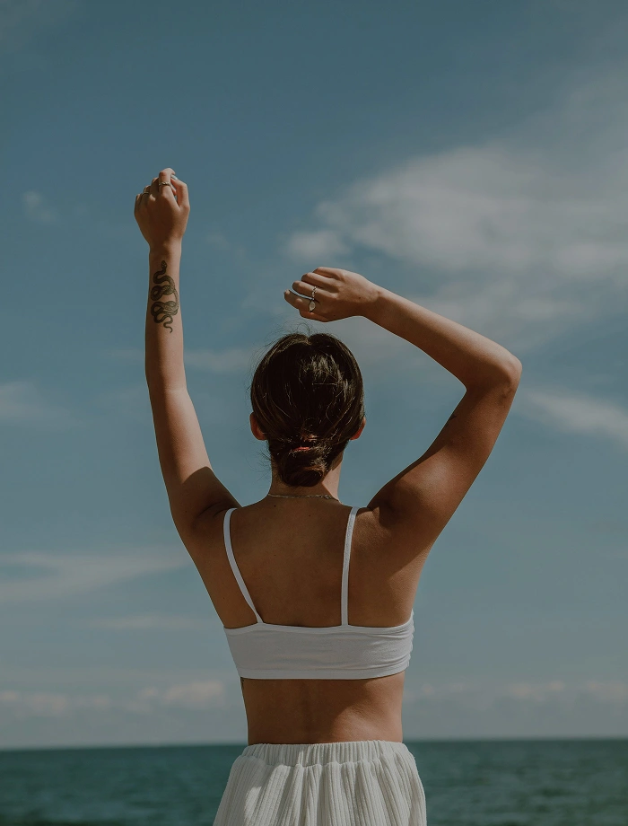 Woman with a snake tattoo on her arm raising both arms against a clear sky near the ocean.