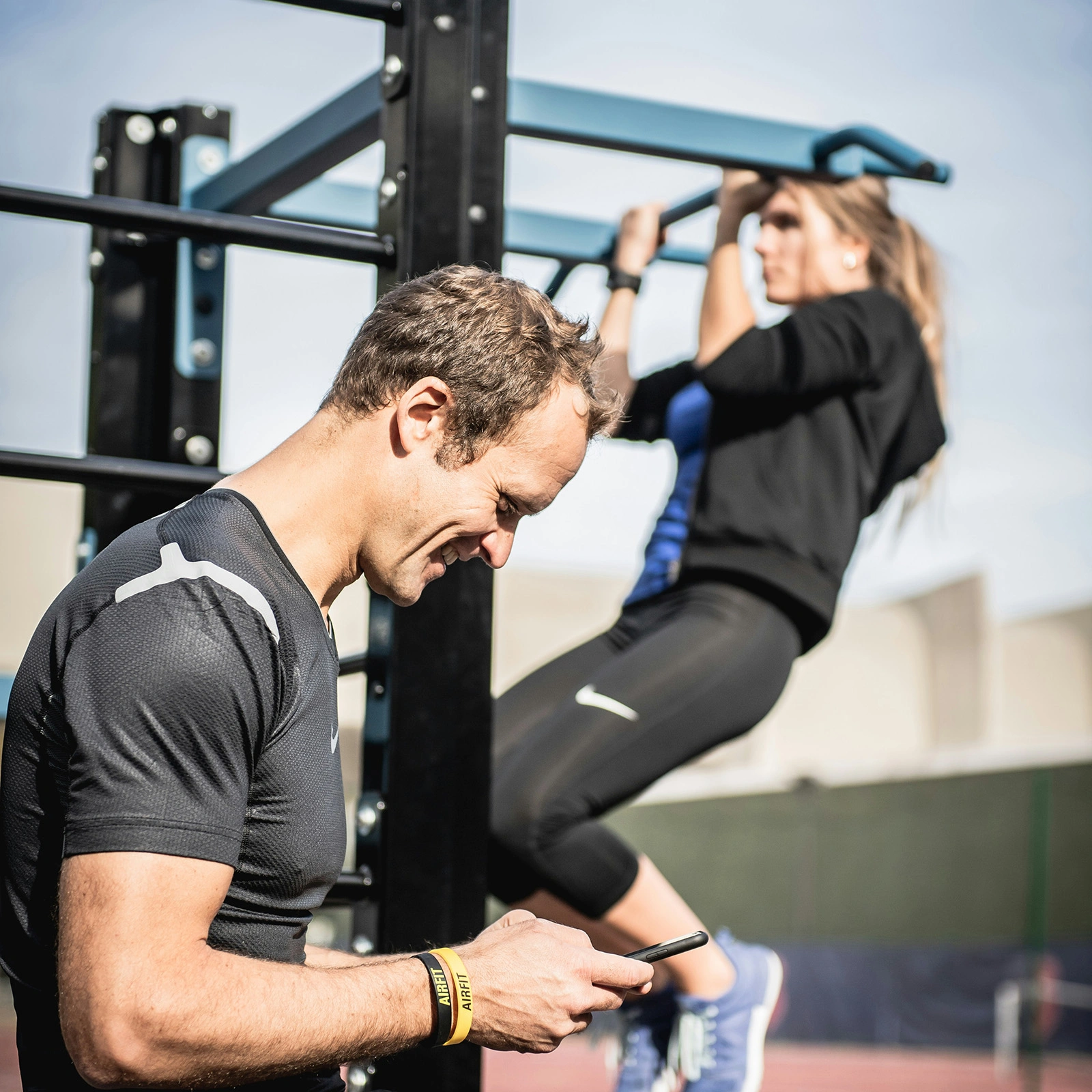 Man in black athletic wear looking at his phone with a woman doing pull-ups on a bar in the background.