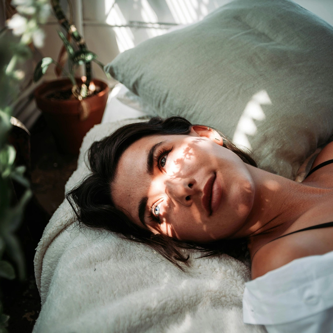 Woman lying on a bed with sunlight and shadows of leaves cast on her face, with a green pillow and a potted plant nearby.