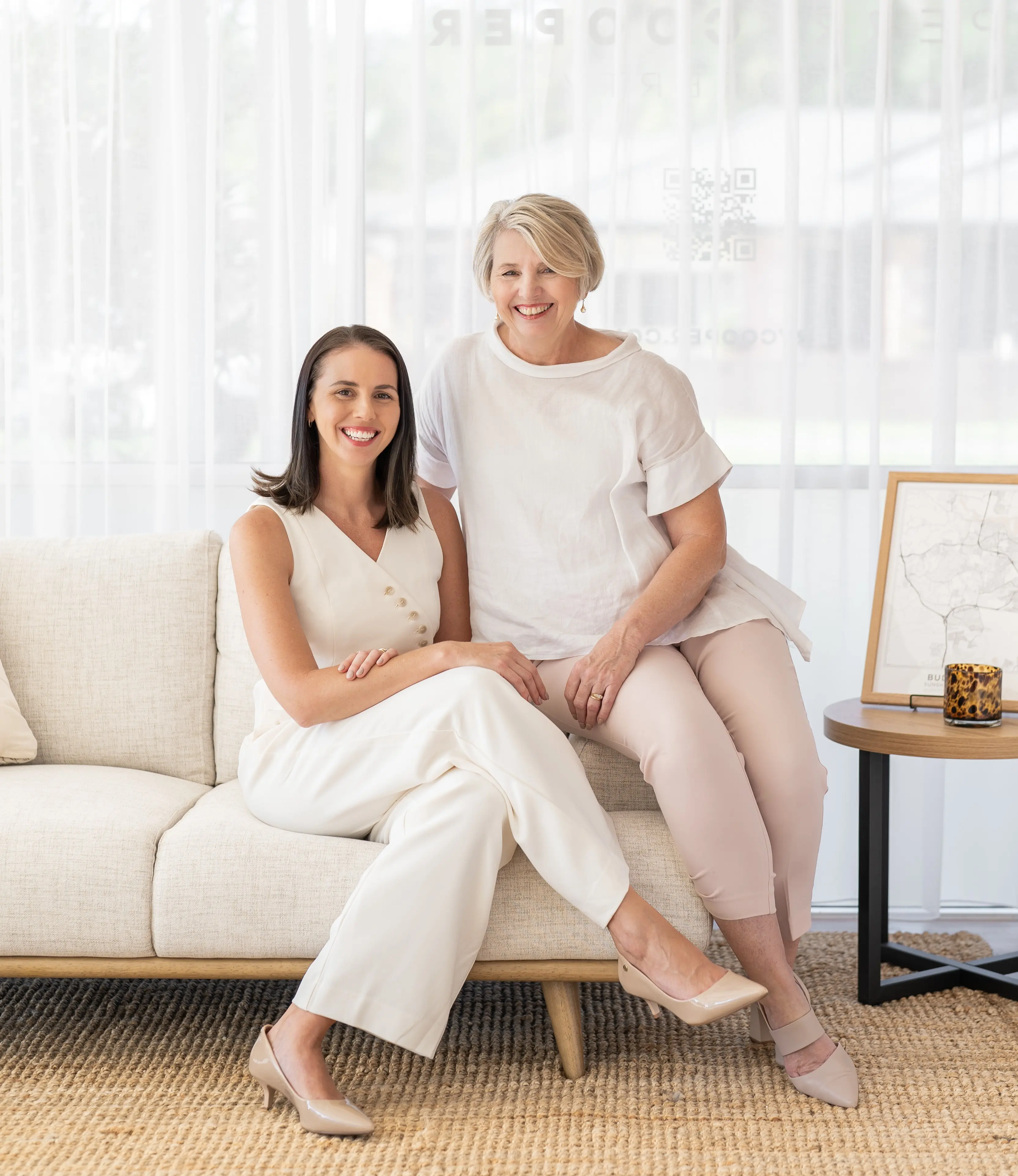 Two smiling women, one seated on a beige sofa and the other sitting on the armrest, dressed in light-colored outfits in a bright, modern room.